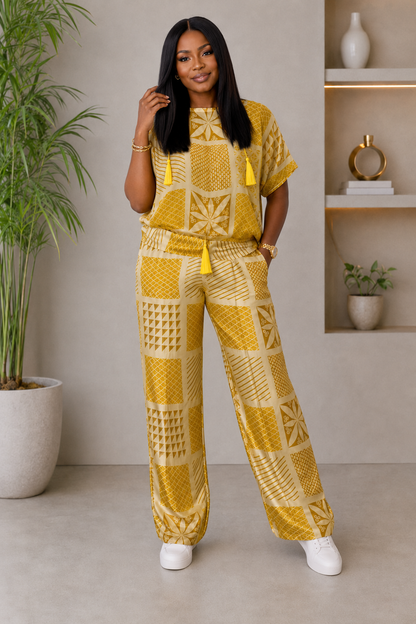 A woman modeling a matching yellow and cream patterned two-piece set consisting of a short-sleeve top and wide-leg pants, styled with white sneakers in a modern indoor setting.