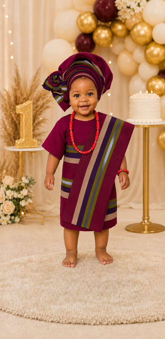 Girl wearing a burgundy striped Aso Oke dress with a matching Yoruba gele headwrap and shoulder sash for a first birthday celebration