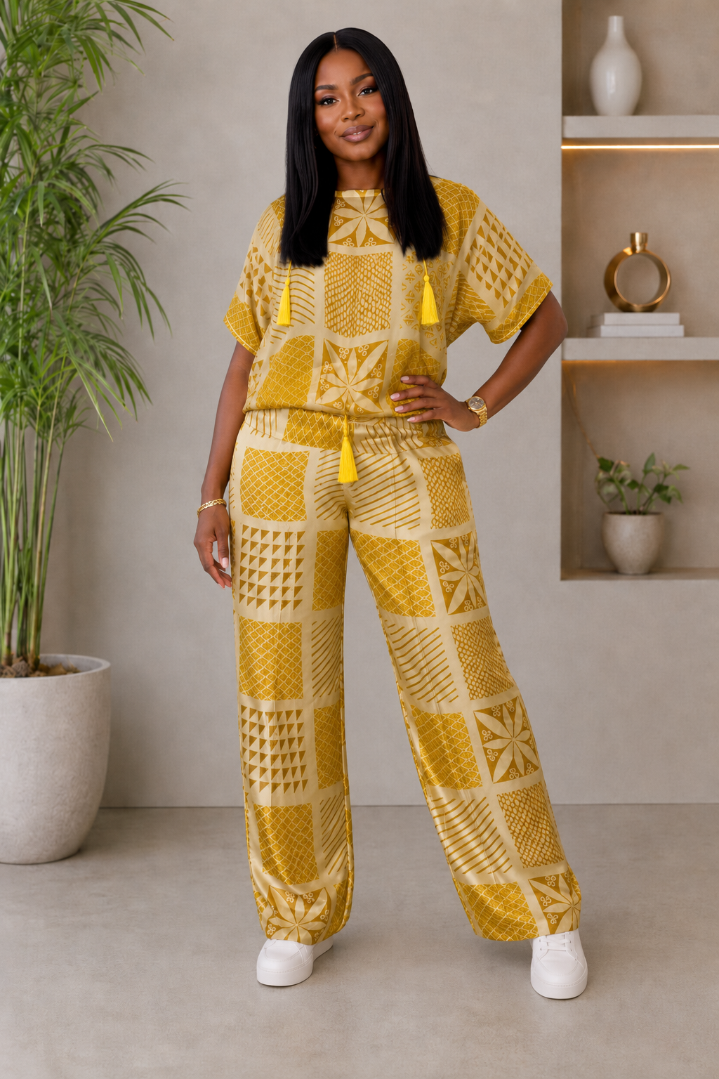 A woman modeling a matching two-piece outfit consisting of a short-sleeve tunic top and wide-leg pants, both in a mustard yellow and cream geometric pattern with yellow tassels, set against an indoor background with a potted plant and shelves.