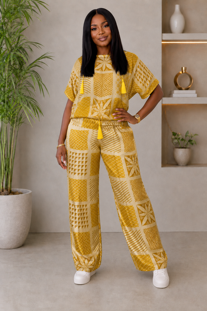 A woman modeling a matching two-piece outfit consisting of a short-sleeve tunic top and wide-leg pants, both in a mustard yellow and cream geometric pattern with yellow tassels, set against an indoor background with a potted plant and shelves.