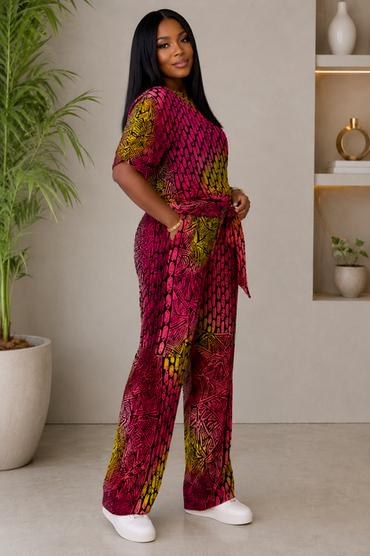 A woman modeling a short-sleeve wrap-style top and pants set with a bold magenta, yellow, and green abstract print, standing in a modern indoor setting with a potted plant and shelving.